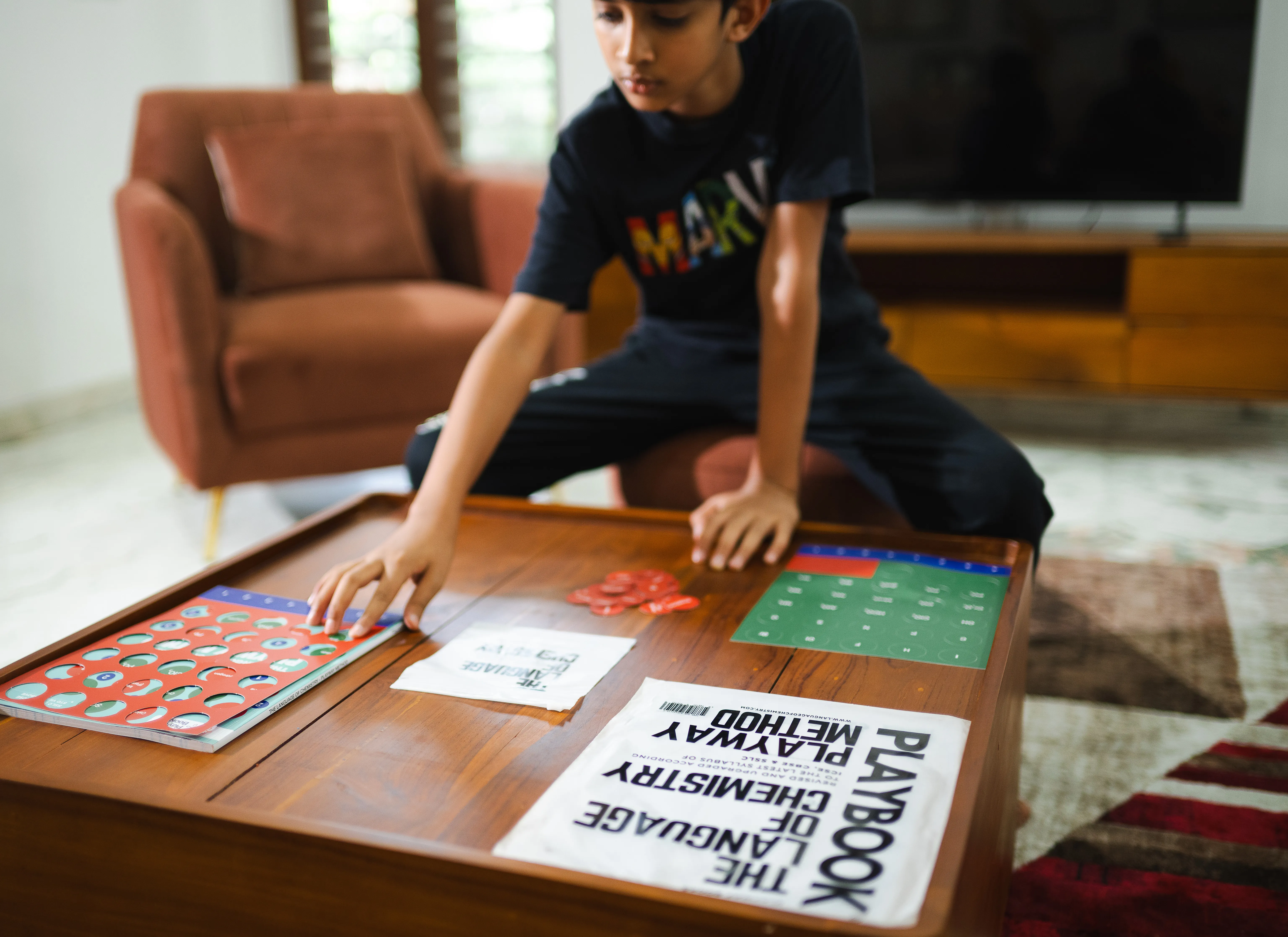 A student reaches across a coffee table spread with the Playbook: the red element sheet and green radical sheet laid flat, a pile of punched-out tokens, and the book's cloth pouch.