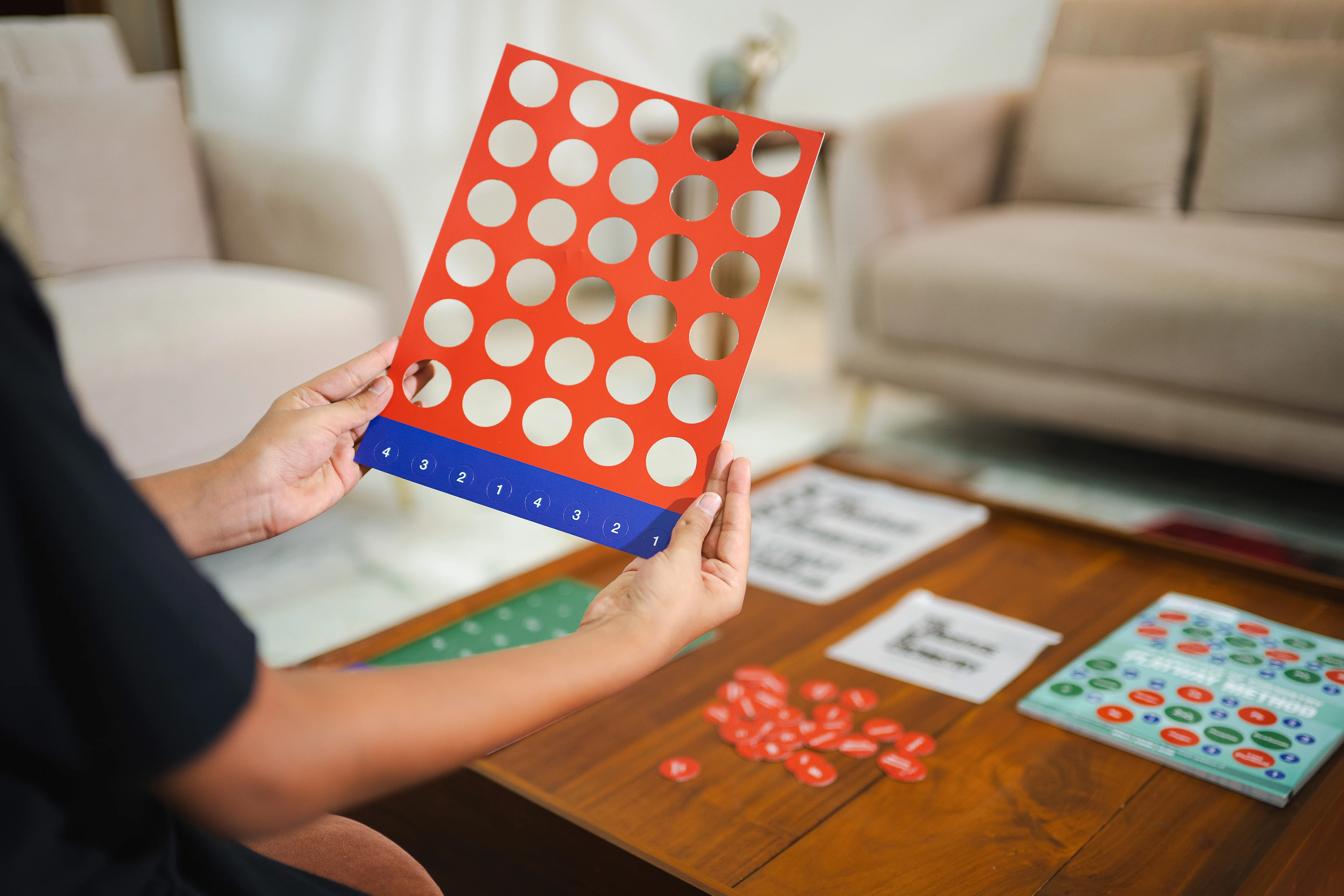 A hand holds up the red element sheet, roughly half its circular tokens already punched out, over a coffee table scattered with the loose tokens that fell free.